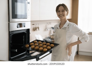 Young attractive woman in apron holding baking tray with fresh baked prepared cupcakes smile look at camera posing stand in modern kitchen near electric oven. Housewife chores, culinary, family recipe - Powered by Shutterstock