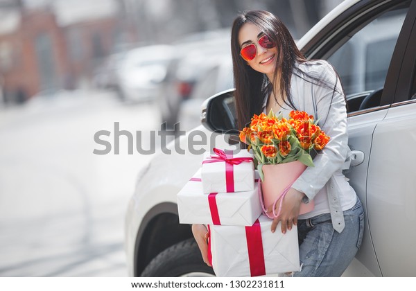Young attractive woman in the car with
presents box, gift and flowers. Beautiful lady in spring time with
bouquet of tulips. Female in the
automobile.