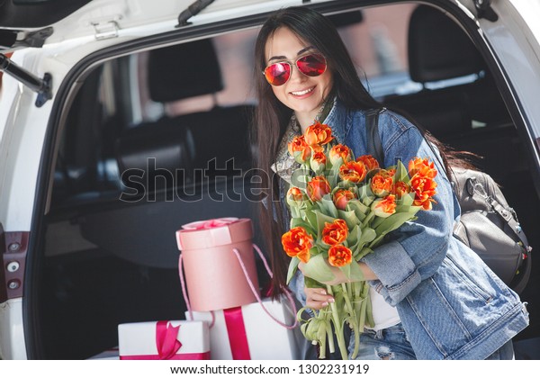 Young attractive woman in the car with\
presents box, gift and flowers. Beautiful lady in spring time with\
bouquet of tulips. Female in the\
automobile.