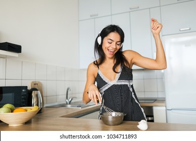 young attractive woman cooking scrambled eggs in kitchen in morning, smiling, happy positive housewife, healthy lifestyle, listening to music on headphones, laughing, having fun, dancing, singing