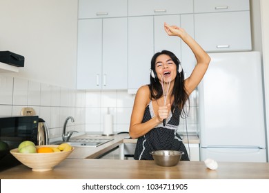 young attractive woman cooking scrambled eggs in kitchen in morning, smiling, happy positive housewife, healthy, listening to music on headphones, singing in whisk like in microphone, having fun