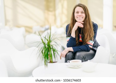 Young Attractive Woman With A Cup Of Tea In A Cafe.  Smiling, Gap Teeth. Bright Modern Interior. Natural Unconventional Beauty. Communication With Riends, Conversation, Talk. Looks At The Camera