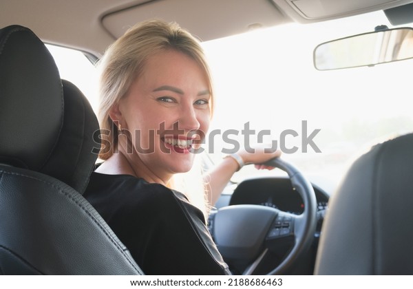 Young attractive\
woman drives a car. Beautiful blonde woman with long hair smiling\
in a car while driving. Woman turned around from the driver\'s seat\
and looks at the camera