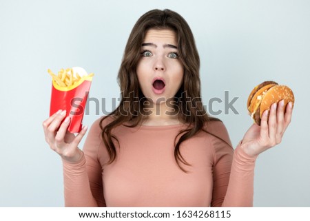 young attractive woman holding meat burger in one hand and fried potatoes in another looking amazed on isolated white background, dietology and nutrition