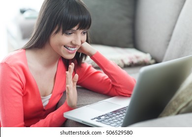 Young attractive woman at home connecting to the internet with a laptop and videocalling, she is waving her hand