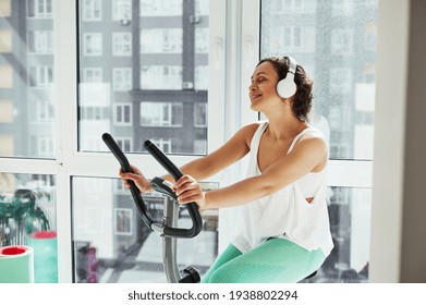 Young Attractive Woman Listen To The Music While Working Out On Spin Bike Against Large Windows At Home. Cardio Training Concept