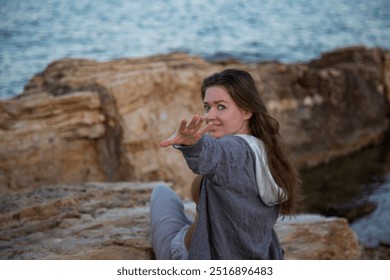 young attractive woman with long brown hair and blue eyes sitting on rocky beach smiling and reaching forward with her hand - Powered by Shutterstock