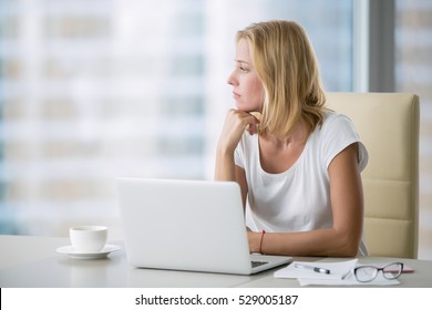 Young attractive woman at a modern office desk, working with laptop, looking at the window, thinking about a post, full-time blogger, seeking for inspiration, help to be productive, updating computer