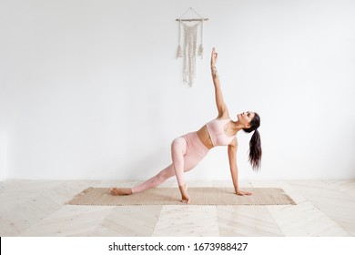 Young attractive woman practicing yoga in bright yoga class on wooden eco floors tanding in Full Version of Vasisthasana exercise, Side Plank pose, working out, pink sportswear. White wall background - Powered by Shutterstock