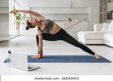 Young Attractive Woman Practicing Yoga And Stretching Body At Home Using Laptop For Online Class Or Virtual Tutorials. Girl Standing In Utthita Parsvakonasana Exercise, Doing Side Angle Pose. Wellness