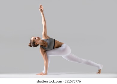 Young attractive woman practicing yoga, standing in Revolved Side Angle exercise, Parivrtta Parsvakonasana pose, working out wearing sportswear, indoor full length, isolated, grey studio background