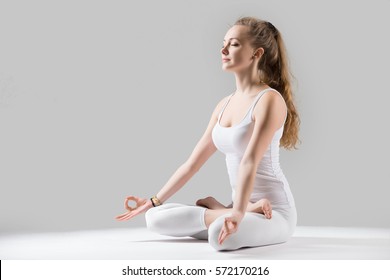 Young attractive woman practicing yoga, sitting in Padmasana exercise, Lotus pose on meditation session, working out wearing sportswear white tank top, full length, isolated, grey studio, closed eyes 