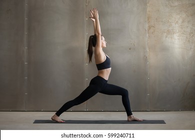 Young attractive woman practicing yoga, standing in Warrior one exercise, Virabhadrasana I pose, working out, wearing black sportswear, cool urban style, full length, grey studio background, side view