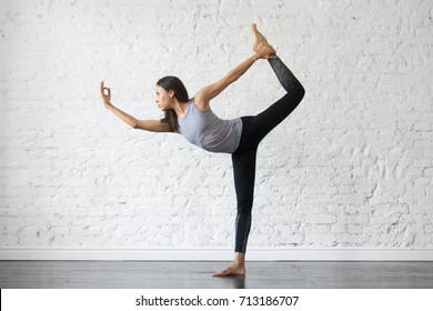 Young attractive woman practicing yoga, stretching in Natarajasana exercise, Lord of the Dance pose, working out, wearing sportswear, gray tank top, black pants, indoor full length, studio background  - Powered by Shutterstock