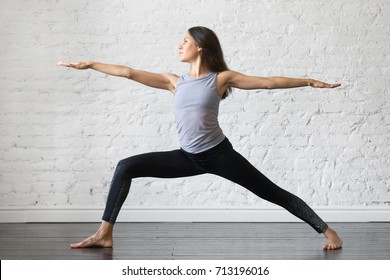 Young Attractive Woman Practicing Yoga, Standing In Warrior One Exercise, Virabhadrasana II Pose, Working Out, Wearing Sportswear, Gray Tank Top, Black Pants, Indoor Full Length, Studio Background 