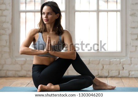 Young attractive woman practicing yoga, doing Half lord of the fishes exercise, Ardha Matsyendrasana pose with namaste , working out, wearing sportswear, pants and top, indoor full length, yoga studio