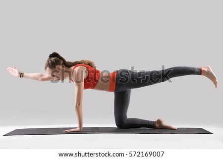 Young attractive woman practicing yoga, standing in Bird dog exercise, Donkey Kick pose, working out, wearing sportswear, red tank top, pants, indoor full length, isolated, grey studio background 