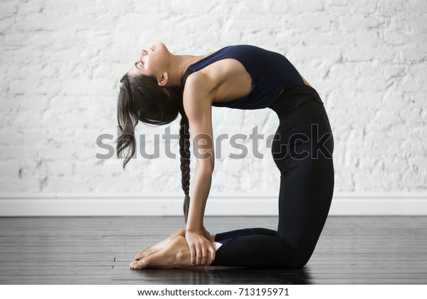 Young attractive woman practicing yoga, stretching in Ustrasana exercise, Camel pose, working out, wearing sportswear, black top and pants, indoor full length, studio background 