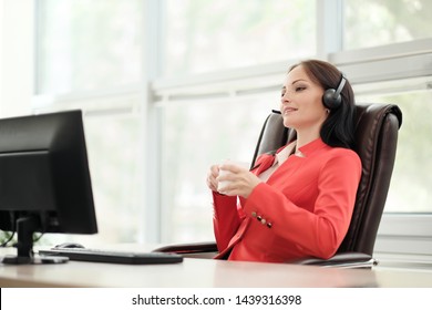Young attractive woman in red jacket sitting on desk at head of executive in red jacket. Holds video conference in headphones with microphone. Drinking coffee from white cup. Smiles and communicates.