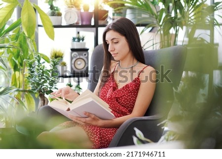 Young attractive woman relaxing at home surrounded by beautiful plants, she is sitting and reading a book
