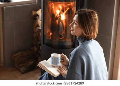 Young attractive woman sitting close to fireplace, reading interesting book and drinking hot tea in the cozy house - Powered by Shutterstock