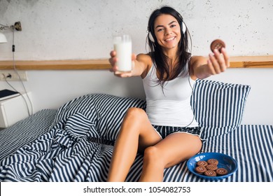 young attractive woman sitting on bed in pajamas having breakfast, smiling, bedroom, happy emotion, wake up in morning, listening to music on headphones, drinking milk, eating cookies, healthy