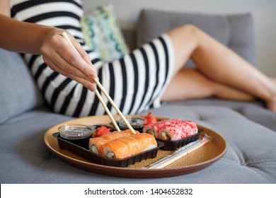 Young attractive woman sitting on the sofa and eating sushi at home. Food delivery concept background