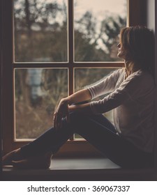 Young attractive woman, sitting on a window, looking outside, lonely mood
