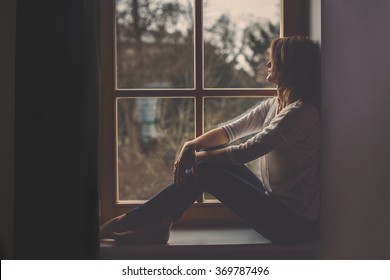 Young attractive woman, sitting on a window, looking outside, lonely mood