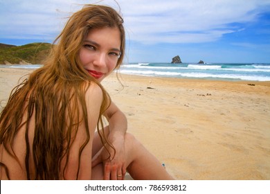 Young attractive woman skinny dipping in paradise wild beach, long hair natural nude girl sitting alone on golden sand during the clear sunny day in front of seascape with ocean waves on background 