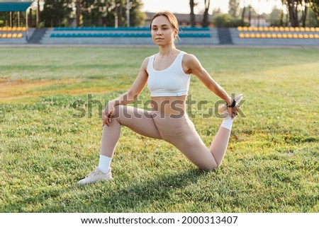 Young attractive woman in sportswear doing pre-training warm-up, legs stretching at the stadium while standing on knee. Healthy lifestyle, outdoor training.