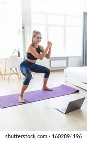 Young attractive woman in sportswear making deep sumo squats in living room while watching fitness online video on laptop. Lady doing exercises for maintaining optimum weight and good physical shape