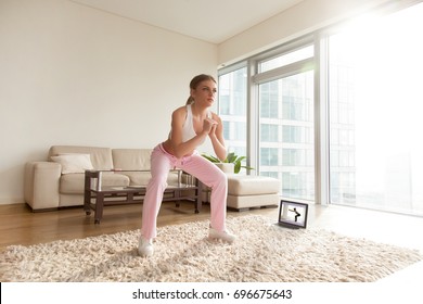 Young attractive woman in sportswear making deep sumo squats in living room while watching fitness online video on laptop. Lady doing exercises for maintaining optimum weight and good physical shape - Powered by Shutterstock