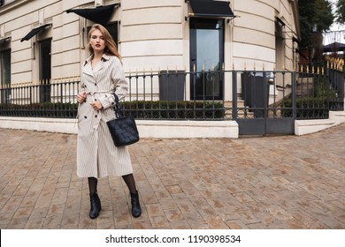 Young attractive woman in striped trench coat with black handbag dreamily looking in camera walking around cozy city street - Shutterstock ID 1190398534