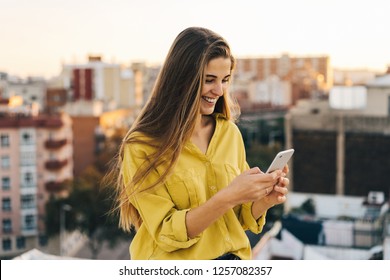 young attractive woman using her smartphone for send a message. female in a yellow blouse smiling on the rooftop while light is magic. Girl checking mobile phone. business woman watching at the screen