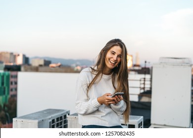 Young Attractive Woman Using Her Smartphone For Make Online Shopping. Female In A White Sweater Smiling. Checking Mobile Phone. Business Woman Looking At The Screen. Girl E-learning Via Her Gadget 