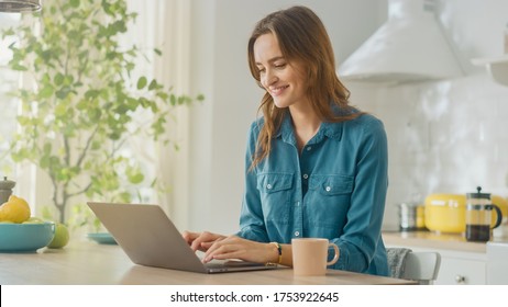 Young Attractive Woman Using Laptop Computer and Drinking Morning Cup of Coffee While Wearing a Jeans Shirt. Brunette Female Sitting in a Modern Cozy Kitchen. Freelancer Working from Home. - Powered by Shutterstock