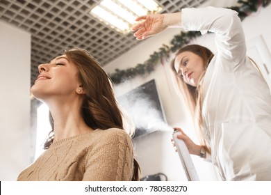 Young attractive woman visiting hairdresser. Woman in white shirt making hairstyle to a woman in beige sweater 