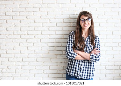 Young attractive woman wearing checkered shirt & cat eye glasses smiling wide. Pretty female model, hands crossed on chest, brunette hair posing. Girl, white brick wall background, copy space, closeup