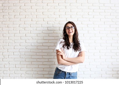 Young attractive woman wearing white shirt & cat eye glasses smiling wide. Pretty female model, hands crossed on chest, brunette hair posing. Girl, white brick wall background, copy space, closeup - Powered by Shutterstock