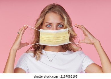 A young attractive woman in a white casual t-shirt wears a protective face mask. Protection against flu and cold diseases during a pandemic. Pink isolated background - Shutterstock ID 2189082195