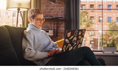 Young Attractive Woman Working from Home on Laptop Computer with Stickers in Sunny Stylish Loft Apartment. Creative Designer Wearing Cozy Blue Sweater and Glasses. Urban City View from Big Window.
