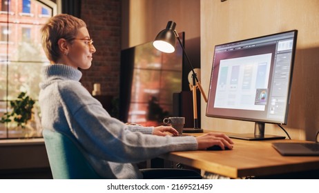 Young Attractive Woman Working from Home on Desktop Computer in Sunny Stylish Loft Apartment. Creative Designer Wearing Cozy Blue Sweater and Glasses. Urban City View from Big Window.