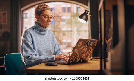 Young Attractive Woman Working from Home on Laptop Computer with Stickers in Sunny Stylish Loft Apartment. Creative Designer Wearing Cozy Blue Sweater and Glasses. Urban City View from Big Window. - Powered by Shutterstock