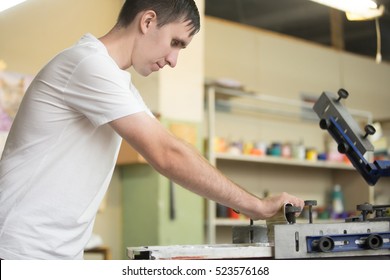 Young Attractive Worker Man Working Using Printmaking Tools, Silk Screen Printing On Clothing Fabric Technique 