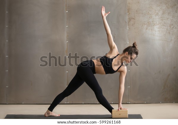 Young attractive yogi woman in black practicing yoga,\
standing in Utthita Trikonasana exercise using block, extended\
triangle pose, working out, cool urban style, full length, grey\
studio background 