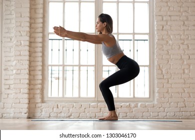 Young Attractive Yogi Woman Practicing Fitness, Doing Bodyweight Squat Exercise, Yoga Chair Pose, Working Out, Wearing Sportswear, Black Pants And Top, Indoor Full Length White Yoga Studio