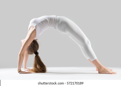 Young attractive yogi woman practicing yoga, standing in Bridge exercise, Urdhva Dhanurasana pose, working out, wearing sportswear, white tank top, pants, full length, isolated, grey studio background