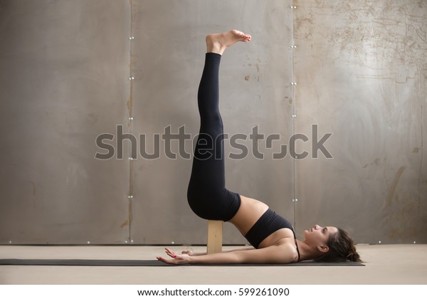 Young attractive yogi woman practicing yoga, standing in\
Viparita Karani exercise, using wooden block, working out, wearing\
black sportswear, cool urban style, full length, grey studio\
background 