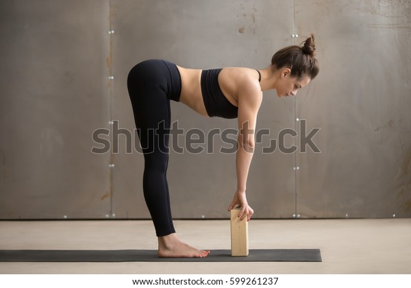 Young attractive yogi woman practicing yoga,\
standing in head to knees, Ardha uttanasana exercise, using block,\
Standing Half forward bend pose, working out, cool urban style,\
full length, grey studio